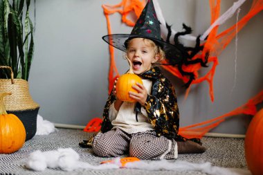 Cadılar Bayramı kostümü giymiş, elinde balkabağı olan ve arka planda şenlikli bir dekorla gülen genç bir kızın neşeli ve samimi bir yakın plan fotoğrafı..