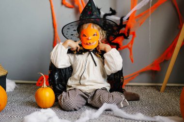 Neşeli bir çocuk, balkabakları ve Cadılar Bayramı dekoru arasında oturur. Üzerinde büyüleyici bir kostüm ve oyulmuş bir balkabağı maskesi vardır. Tatili kutlarken..