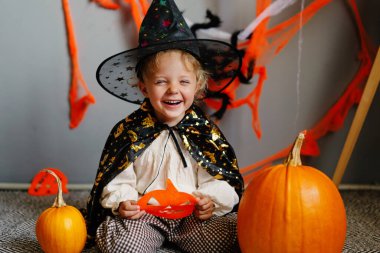 Cadı kostümü giymiş neşeli bir çocuk canlı süslemeler ve balkabaklarıyla çevrili bir balkabağı maskesi tutuyor. Cadılar Bayramı 'nı neşeyle kutluyor..