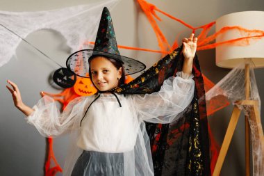 Genç bir cadı örümcek ağları ve Cadılar Bayramı ışıklarıyla dolu bir odada mutlu bir şekilde poz veriyor. Kutlama yaparken büyülü bir kostüm giyiyor..