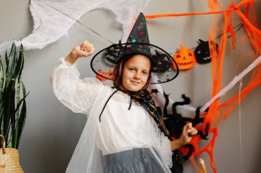 Cadı gibi giyinmiş genç bir kız Cadılar Bayramı için dekore edilmiş bir yerde mutlu bir şekilde poz veriyor. Etrafı ürkütücü süslemeler ve neşeli renklerle çevrili..