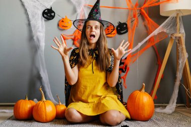 Cadı şapkası ve peleriniyle bir kız, balkabaklarıyla çevrili bir şekilde yerde oturuyor ve Cadılar Bayramı 'nı süslemelerle kutluyor..