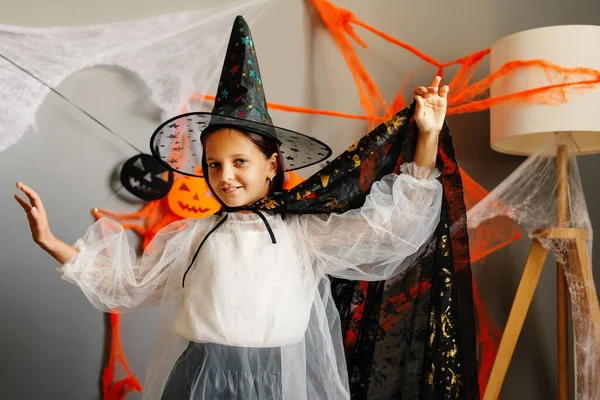 Genç bir cadı örümcek ağları ve Cadılar Bayramı ışıklarıyla dolu bir odada mutlu bir şekilde poz veriyor. Kutlama yaparken büyülü bir kostüm giyiyor..