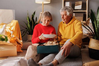 Büyüleyici bir oturma odasında, yaşlı bir çift, canlı yeşillik ve yumuşak ışıkla çevrili bir hediyeyi birlikte keşfederken mutluluğu paylaşıyor..