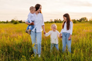 Bir anne, bebeğini kucağında tutarken gülümserken iki küçük kızı da birbirine uyumlu beyaz gömlekler ve kotlar giyip güneşli bir çayırda gülüyor ve onunla yürüyor..