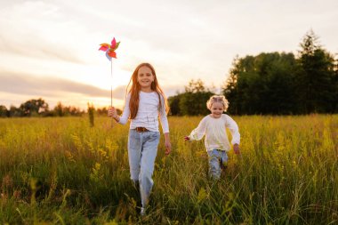 İki neşeli genç kız kardeş gün batımında güneşli bir tarlada koşarken yakalandı. Büyük kız kardeş renkli bir fırıldak tutarken, küçük olan gülümseyerek onun yanında mutlu bir şekilde koşuyor..
