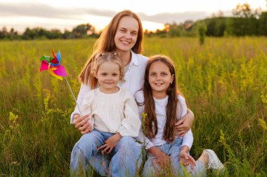 Gün batımında bir tarlada oturan gülümseyen bir anne ve iki küçük kızının güzel bir portresi. En küçük kızın elinde renkli bir fırıldak var..