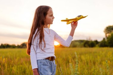 Güneşli bir alanda elinde sarı oyuncak bir uçakla duran genç bir kız gün batımında onu altın gökyüzüne fırlatmaya hazırlanıyormuş gibi bakıyordu..