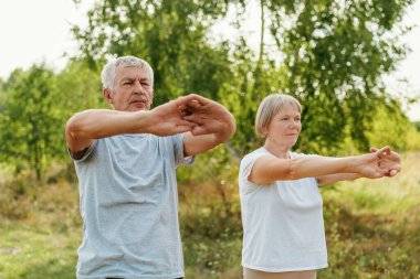 Yaşlı bir çift kollarını uzatır ve güneşli bir sabahta huzurlu bir parkta doğanın güzelliğinin tadını çıkararak sağlık ve sağlığı desteklerler..