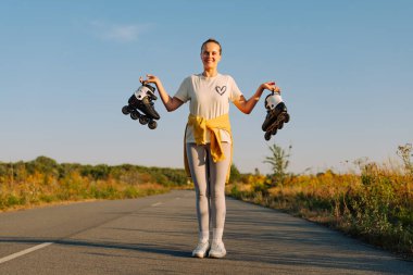 Genç bir kadın kaldırımlı bir yolda elinde patenlerle, arkasında canlı bir manzarayla, öğleden sonra güneşiyle mutlu bir şekilde duruyor..