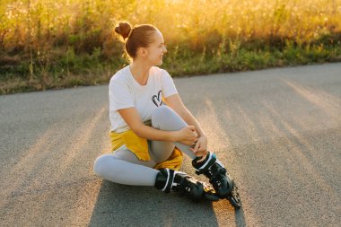 Neşeli bir kız, gün batımında paten kaymaya ara verip, uzun otlar ve sıcak ışıklarla çevrili bir yolda bacak bacak bacak üstüne atarak oturur..
