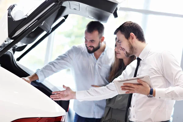 Fiducioso giovane venditore spiegando caratteristiche auto ai giovani proprietari attraenti — Foto Stock