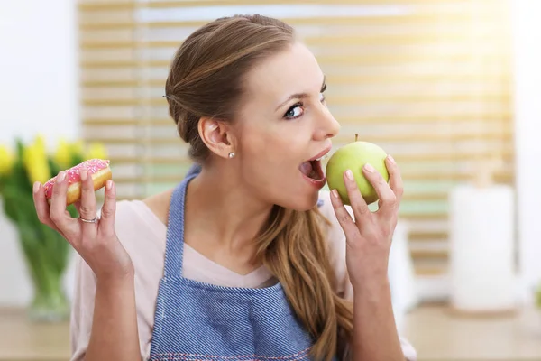 Mulher adulta escolhendo entre maçã e donut na cozinha — Fotografia de Stock