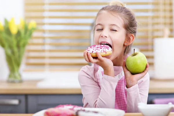 Симпатична дівчинка вибирає між яблуком і пончиком на кухні — стокове фото