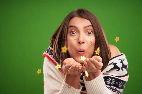 Atractiva morena soplando confeti sobre fondo verde —  Fotos de Stock