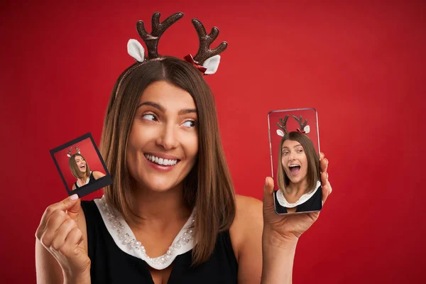 Vrouw in kerstsfeer het vergelijken van Instant Foto's naar smartphone camera over rood — Stockfoto