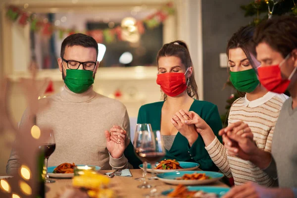 Группа друзей в масках, молящихся за рождественским праздничным столом дома — стоковое фото