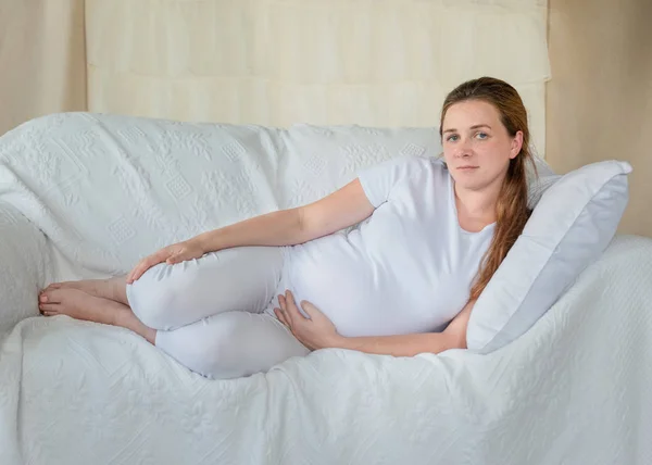 young pregnant woman in home resting on white sofa, embracing her belly, swollen legs