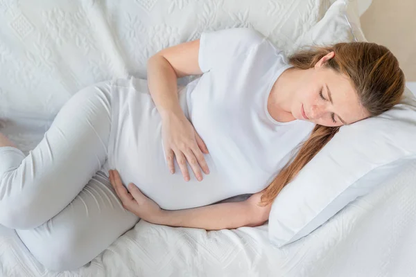 年轻的孕妇在家休息在白色沙发上 拥抱她的肚子 没有化妆 免版税图库图片