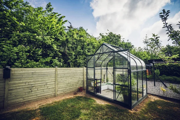 Greenhouse in a garden with trees behind a wooden fence under a blue sky