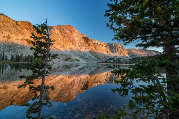 The light of sunrise strikes the sheer rock walls of the Snowy Range in Wyoming, reflecting off the still surface of a lake.