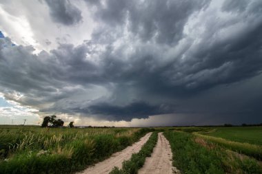 A supercell thunderstorm looms in the sky over a dirt road in farm country. clipart