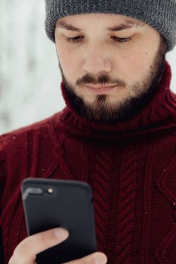 adam kırmızı örgü kazak ve kış ormanın içinde kar üzerinde duran bir telefon tutarak kot