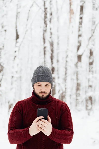 guy in a red knitted sweater and jeans holding a phone, standing on the snow in the winter forest