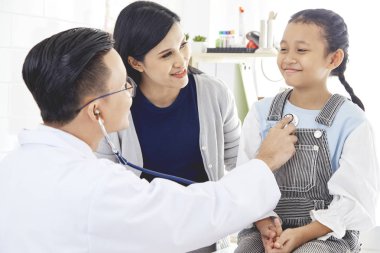 Doktor steteskoptaki hasta bir çocuğun akciğerlerini dinliyor. Annesiyle birlikte doktora geldi. Kız soğuk algınlığından hasta. Doktorun ışık odasındalar..