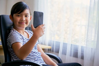 Asyalı küçük kız akıllı telefonu evdeki bir sandalyede video görüşmesi için kullanıyor.
