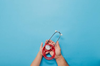 Yetişkin ve çocuk eller kırmızı steteskopu mavi arkaplanda tutuyor ve metin için fotokopi alanı var. Üst görünüm, Sağlık Hizmetleri Tıbbi Konsepti