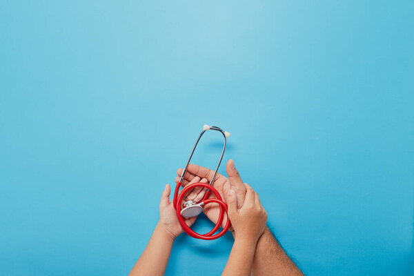 Adult and child hands holding red stethoscope on blue background with copy space for text. Top view, Healthcare medical concept