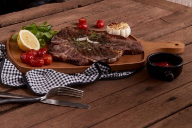 Orta pişmiş ızgara T-Bone Bifteği, otlar, domatesler, limonlar ve sarımsak ve çatal ve bıçakla birlikte kesme tahtasının üzerinde tuz.