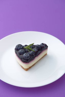 Beyaz tabakta mor arka planda yaban mersinli cheesecake.