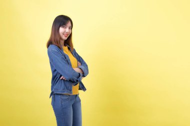 Stüdyoda sarı arka planda genç, şirin ve atletik Taylandlı bir kızın portresi. Çapraz kollu bir kadın sarı tişört, kot ceket ve basit kot pantolon giyiyor.