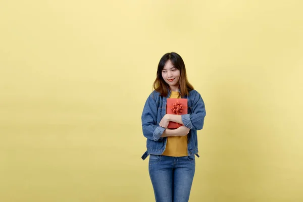 一个年轻的 可爱的和体育的泰国亚洲女孩的肖像在工作室与一个普通的黄色背景 女人抱着礼品盒 穿着黄色T恤牛仔夹克和简单的牛仔裤 — 图库照片