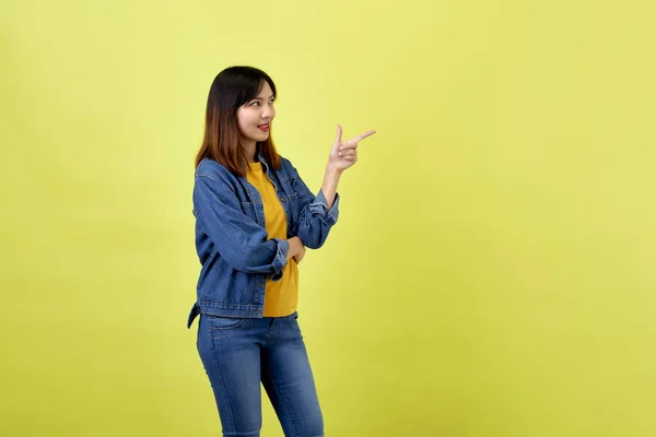 幸せなアジアの女性でデニムジャケットポインティング側へコピースペースとカメラを見ます上の黄色の背景 — ストック写真