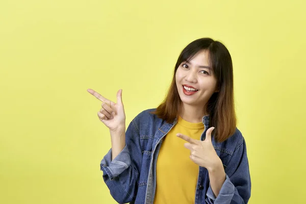 幸せなアジアの女性でデニムジャケットポインティング側へコピースペースとカメラを見ます上の黄色の背景 — ストック写真