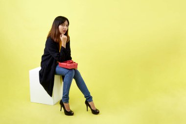 Boş bir kutunun üzerinde oturan mutlu ve çekici genç kadın sarı arka planda kırmızı hediye kutusu tutuyor.