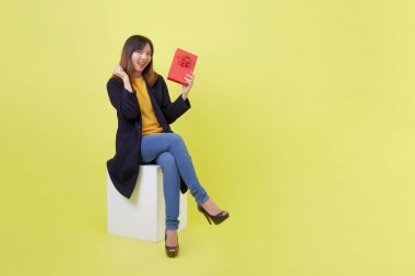 Boş bir kutunun üzerinde oturan mutlu ve çekici genç kadın sarı arka planda kırmızı hediye kutusu tutuyor.