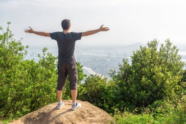 Eller yükselterek ve güzel görünümü ve yeşil çalılar arasında dağın zirvesinde temiz hava keyfini bir adam. Bir kıyı kenti arka planda görünür.