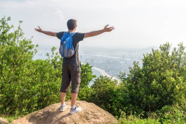 Eller yükselterek ve güzel görünümü ve yeşil çalılar arasında dağın zirvesinde temiz hava zevk turist. Mavi sırt çantası olan adam. Bir kıyı kenti arka planda görünür.