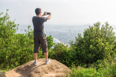 Adam ile fotoğraf çekmek ve güzel görünümü ve yeşil çalılar arasında dağın zirvesinde temiz hava zevk smartphone. Bir kıyı kenti arka planda görünür.