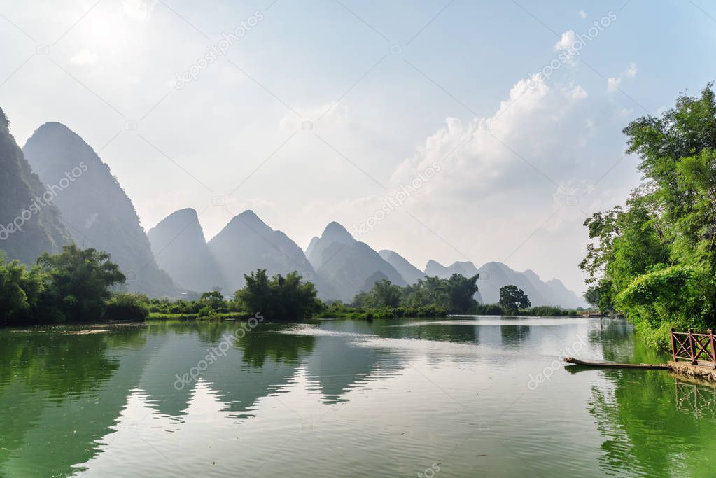 Scenic view of the Yulong River and beautiful karst mountains at Yangshuo County of Guilin, China. Wonderful green hills reflected in water. Small bamboo raft is visible by a pier at right side.