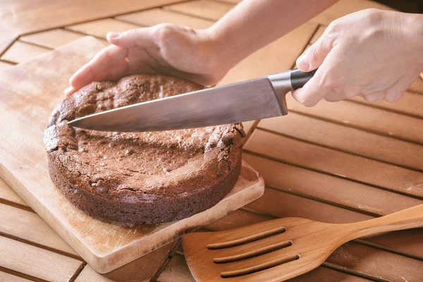 Hand holding knife and cutting delicious freshly baked chocolate brownie cake on wooden table. Homemade sweet dessert, family meal.