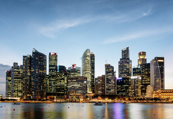 Wonderful evening view of downtown in Singapore. Fantastic skyscrapers and other modern buildings are visible on blue sky background. Amazing colorful city lights reflected in water of Marina Bay.
