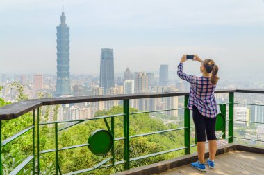 Genç kadın fotoğraf çekiyor ve dağın tepesinden Taipei manzarasının tadını çıkarıyor. Muhteşem Taipei silueti, Tayvan. Harika bir şehir manzarası. Gökdelenler ve şehir merkezindeki diğer modern binalar.