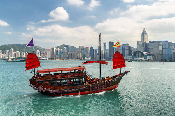 Hong Kong - October 20, 2017: Wonderful view of traditional Chinese wooden sailing ship with red sails in Victoria harbor. Hong Kong Island skyline on sunny day. Amazing cityscape.