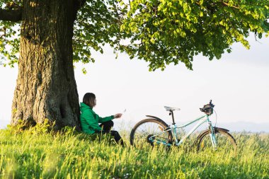 Yalnız bir kız bisikletin yanında büyük yaşlı bir ağacın altında oturuyor.
