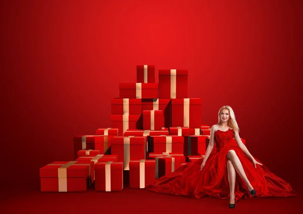 年轻女子坐在红色背景前的礼品盒堆上的销售理念 — 图库照片